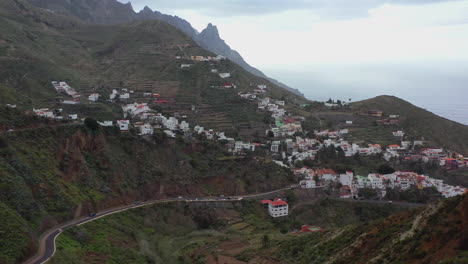 Small-mountain-village-on-the-steep-mountainside-of-Tenerife,-Canary-Islands,-Spain,-sharp-mountain-peaks-above,-coast-of-the-Atlantic-ocean-below-and-stormy-clouds-on-the-horizon,-aerial-4K-shot