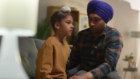 father tying turban onto head of young sikh son with top knot sitting on sofa at home
