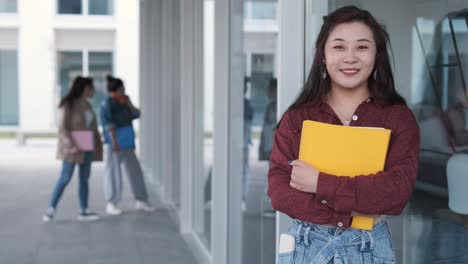 female university student holding folders and looking at the camera while standing at the hallway. education concept.