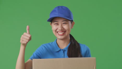 close up of asian female courier in blue uniform showing thumbs up gesture and smiling while delivering a carton on green screen background in the studio