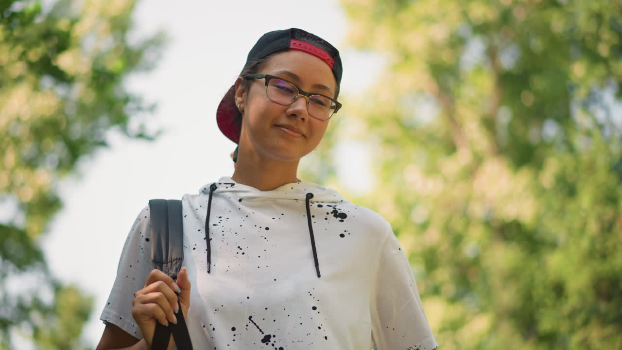 안경과 뒤로 쓴 모자를 착용하고 나무를 올려다보는 젊은 사람, 은은한 미소, 나뭇잎 사이로 비치는 햇빛, 어깨에 백팩 끈, 신선한 공기, 희망에 찬 사색적인 분위기