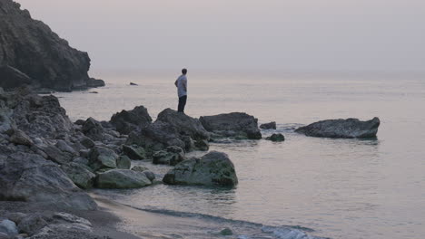 lone man steps onto rock stretching his arms above his head looking at sea as sun rises above horizon