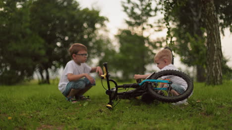 two young siblings are squatting in a grassy field, the younger child plays with the bicycle pedal, while the older brother reacts by leaping up to stop him