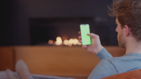 joven relajándose en casa sentado junto al fuego y mirando el teléfono móvil de pantalla verde