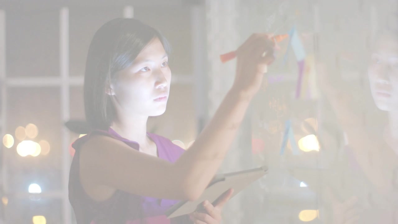Mujer consultando una tablet y organizando notas de colores en una partición de cristal para la planificación empresarial.