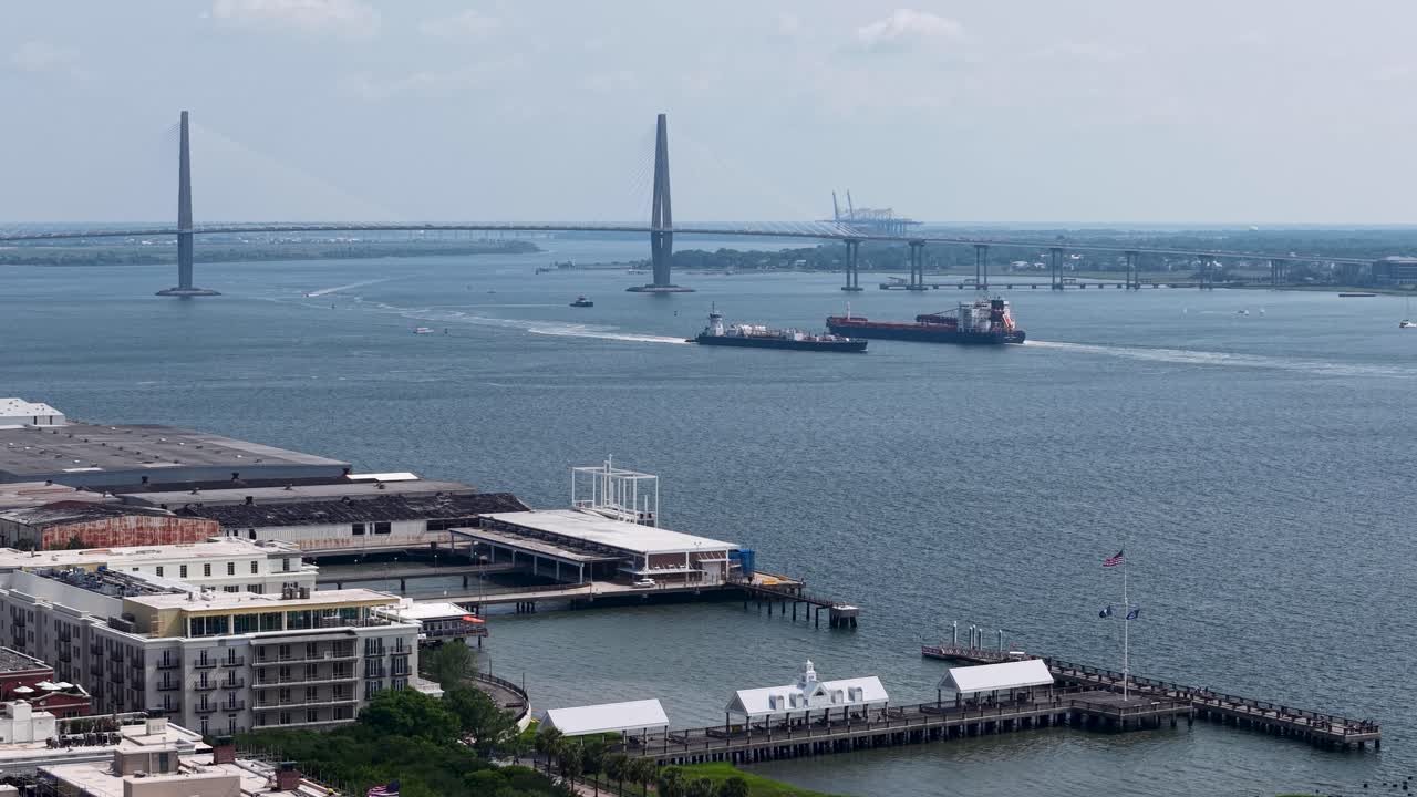 Drone footage shows a cargo ship passing beneath the Arthur Ravenel Jr bridge in Charleston South Carolina. The aerial view captures the Cooper River harbor and waterfront with blue sky