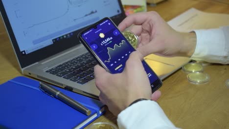 young businessman in his office checking bitcoin chart on his phone while holding physical golden bitcoin in his hands