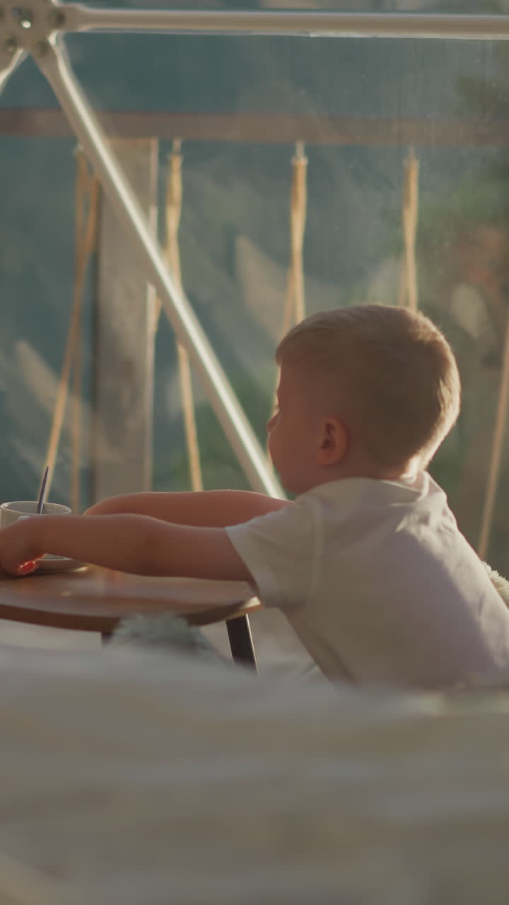 Children sits at table by window. Boy pushes away empty cup. Siblings enjoy peaceful moment in cozy tent at glamping site. Kids on hotel ground