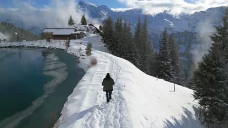 Running-along-the-side-of-the-lake,-a-man-is-headed-to-a-log-cabin-on-the-other-side,-located-on-a-mountain-at-Engelberg,-Brunni,-in-Bahnen,-Switzerland