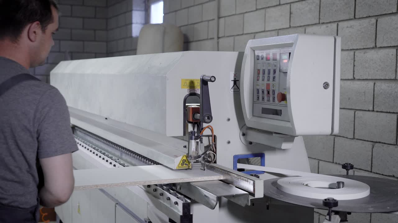 주인은 가구 공장의 작업실에서 파티클 보드의 가장자리를 가공하기 위해 산업 기계를 사용하고 있습니다.