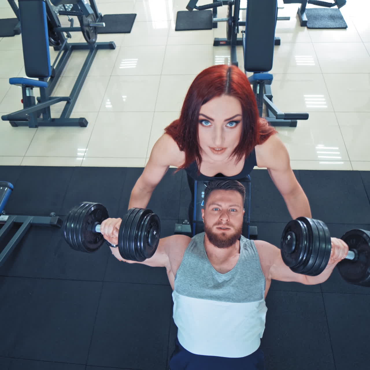 Muscular bearded man is lifting heavy dumbbells on the gym background. Attractive female instructor helps a strong athlete to lift dumbbells. Top view.