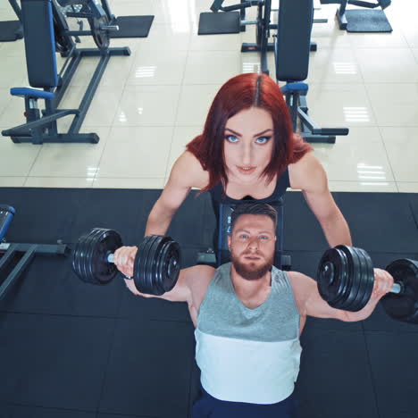 muscular bearded man is lifting heavy dumbbells on the gym background. attractive female instructor helps a strong athlete to lift dumbbells. top view.