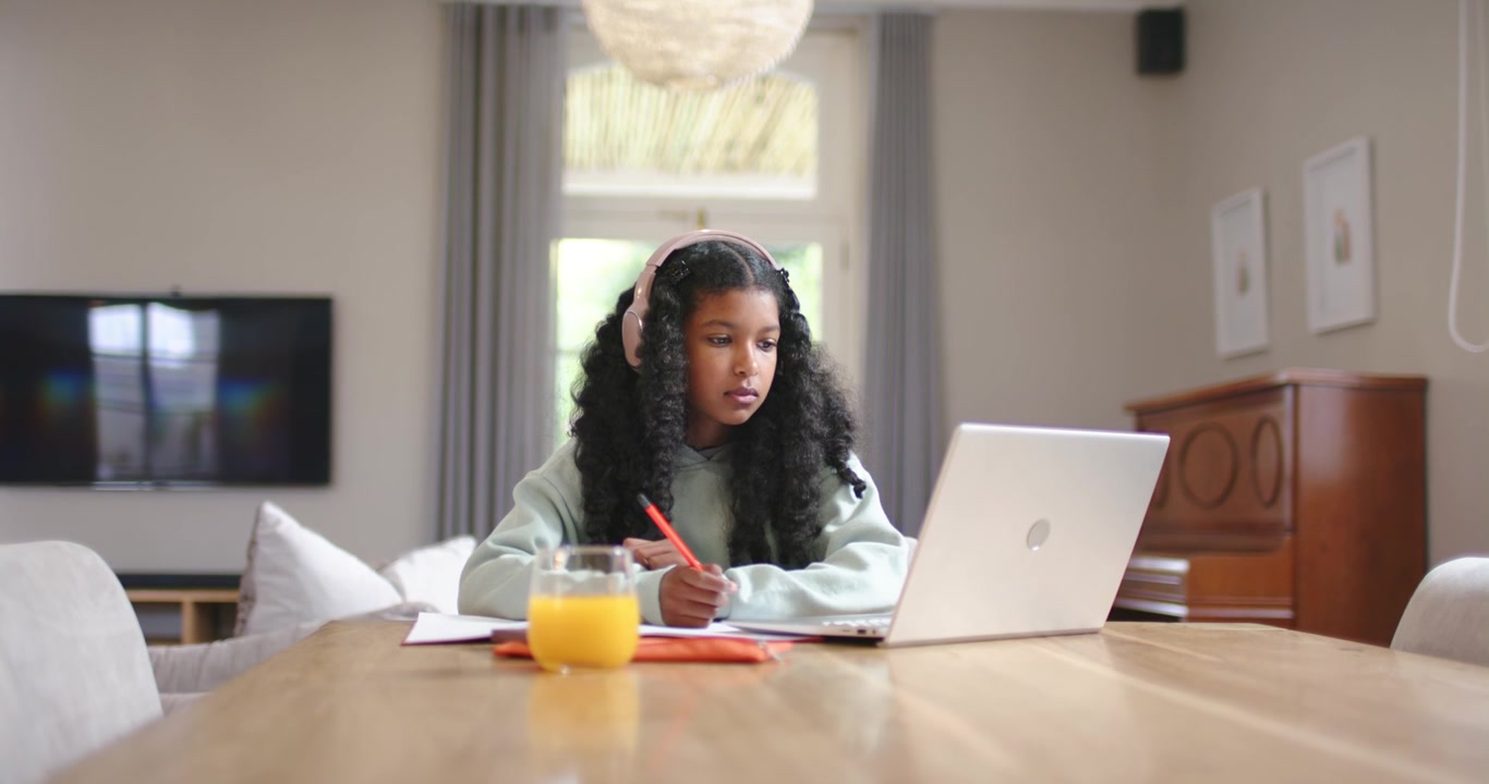 African American teen girl following laptop, taking red-pen notes and inspecting object, studying