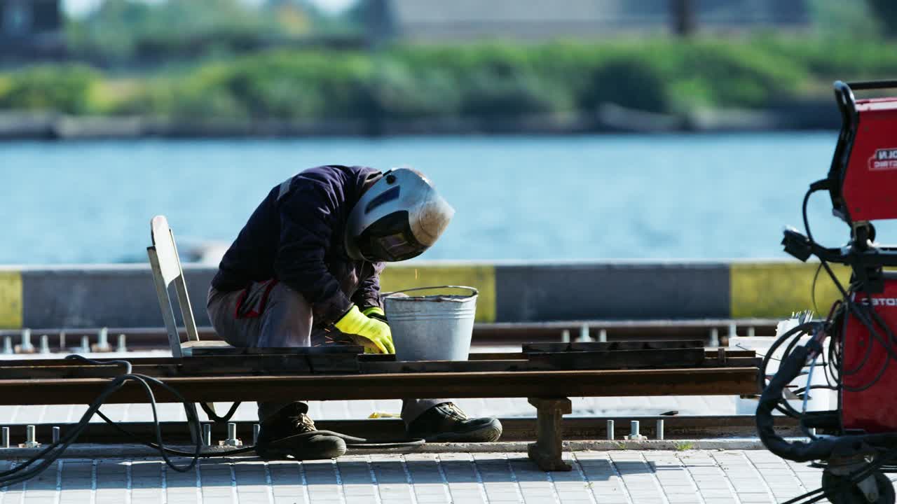 Industrial worker welds metal tracks near waterfront with sparks in calm setting