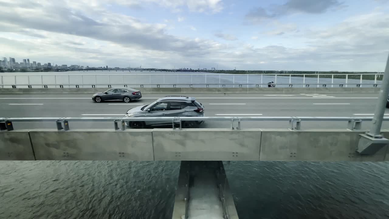 Dynamic view from train passenger perspective of bridge over St. Lawrence River in Montreal, with vehicles in motion, the water below, and the city skyline under a partly cloudy sky, Canada