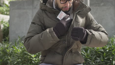 el hombre de etnia hispana se sienta en el banco, revisa una pila de dinero en efectivo en sus manos, luego lo pone dentro de la chaqueta y se va.