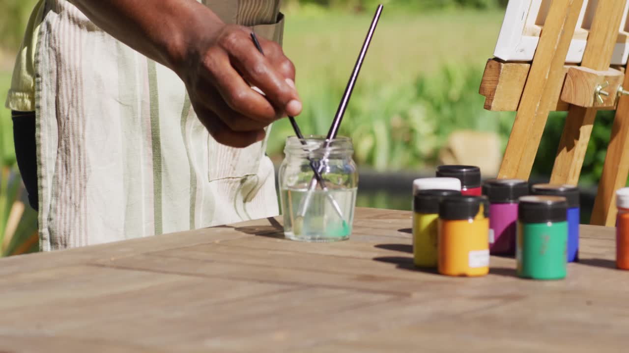 Midsection of african american man painting picture on a canvas in sunny garden cleaning brush