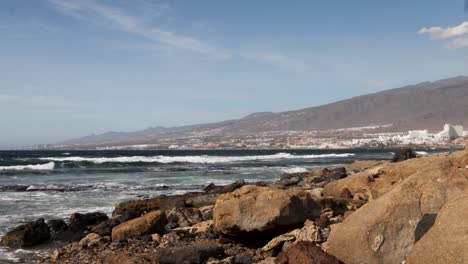 View-of-the-Atlantic-waves-coming-to-the-rocky-shore-with-a-city-in-the-background,-Tenerife