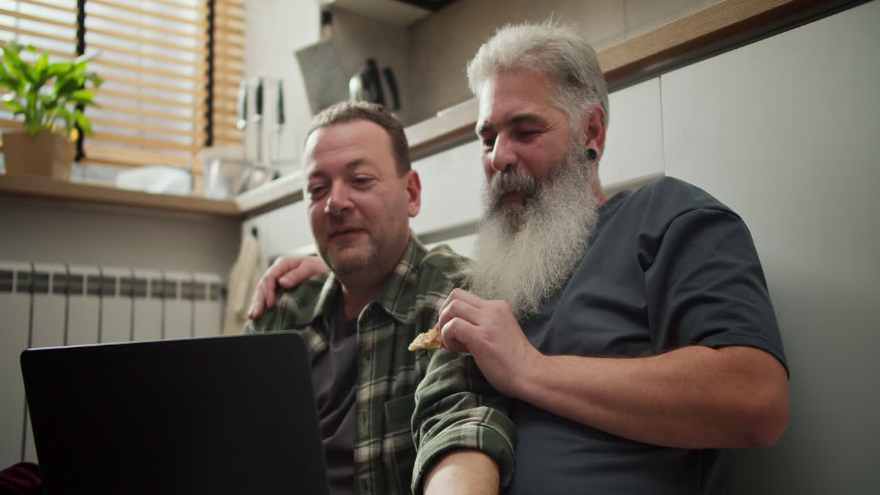 A happy middle-aged man with black hair and stubble together with his elderly boyfriend with a full gray beard sits on the floor in the kitchen eats pizza and looks at something on his gray laptop. During lunch in the afternoon