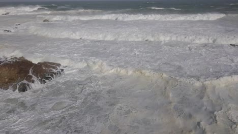 Aerial:-Rows-of-ocean-waves-break-into-whitewater-on-shoreline-rocks