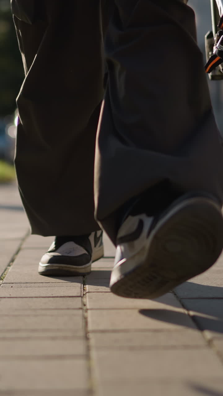 Close-up of legs walking on light gray paved sidewalk wearing loose dark pants and dark sneakers with white soles, with blurred background for focus on movement