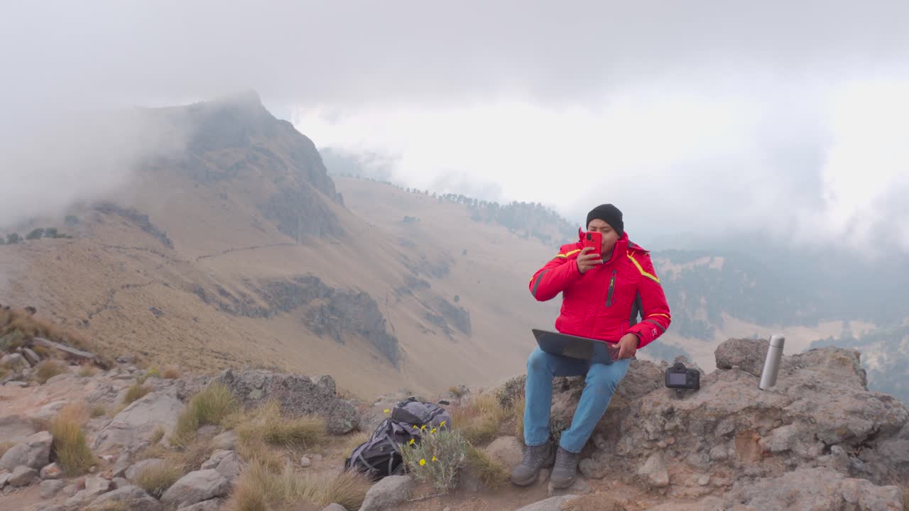 joven latino que trabaja con una computadora portátil en la roca con vistas panorámicas a la montaña, nómada digital en el concepto de viaje
