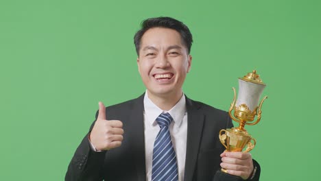 close up of asian business man in a suit and tie with a gold medal and trophy showing thumbs up gesture and smiling to camera as the first winner on green screen background in the studio