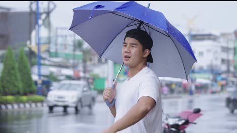 young sporty asian man holding umbrella standing on the street side on the rainy day, unsatisfied stuck in the rain, unlucky day while going out, asia tropical climate season changes, pouring rain