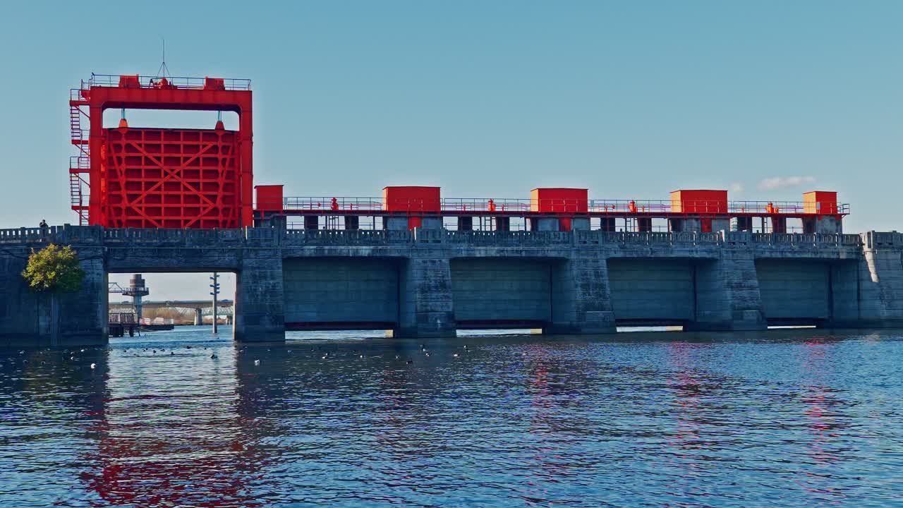 A wide profile shot of the historic Red Sluice Gate. Features a symmetrical view of the industrial steel gates over the river under a clear sky