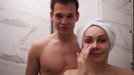 Happy-positive-young-couple-man-and-woman-having-fun-while-standing-in-a-bathroom-after-shower.-Both-playfully-applying-white-mask-on-face.-Having-fun.-Portrait.-Slow-motion