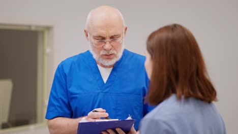 A-confident-man-with-glasses-an-elderly-doctor-in-a-blue-uniform-holds-a-blue-tablet-in-his-hands-and-talks-with-a-brunette-girl-about-her-problems-of-disease-prevention-and-diagnoses-in-a-modern-clinic