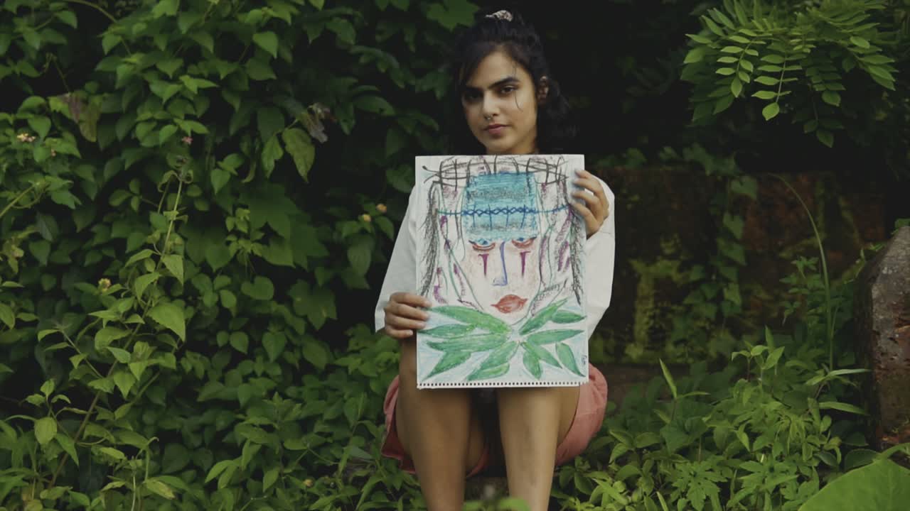 An beautiful girl sitting between plants showing a drawing with an engaging stare