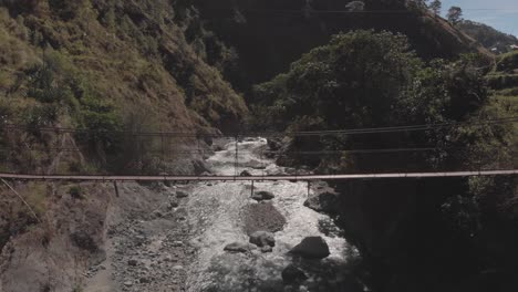 Metal-wire-suspension-bridge-spanning-over-river-in-remote-wilderness-terrain-stationary-medium-wide-angle-aerial