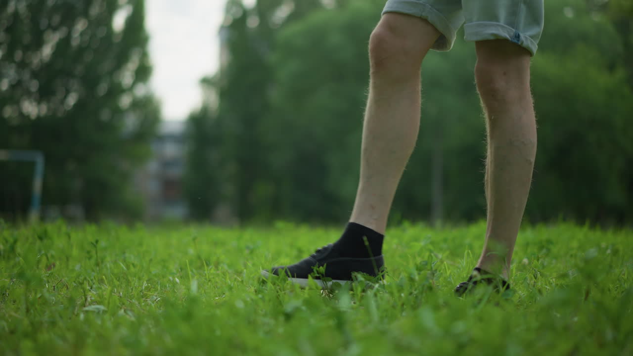A close-up leg view of someone in gray shorts skillfully tapping a soccer ball on a lush, grassy field, with trees in the background