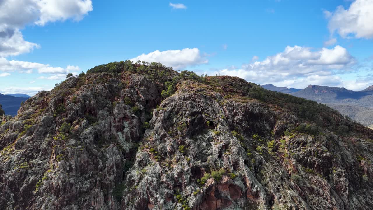 Drone camera smoothly pans over rugged Split Rock, revealing expansive mountain landscape under bright daylight with scattered clouds in Coonabarabran, New South Wales