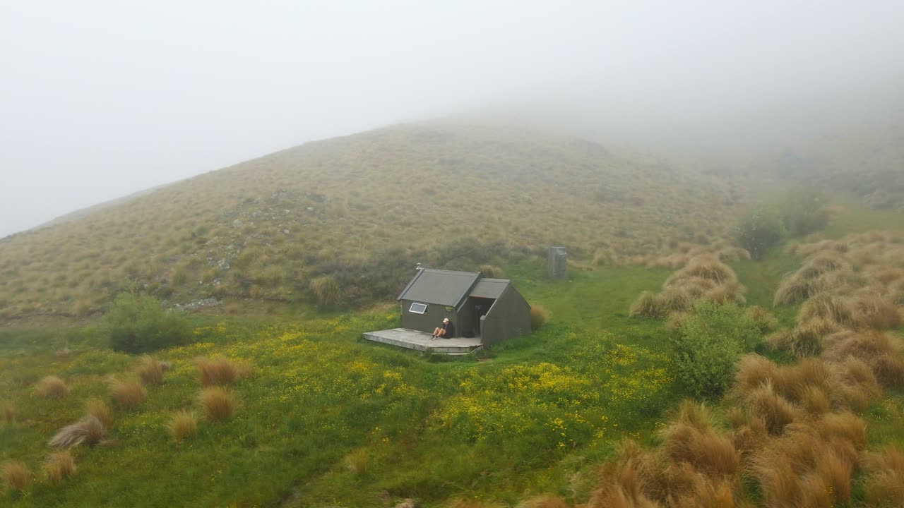 뉴질랜드의 언덕으로 뒤덮인 덤불 속 오두막의 갑판에서 휴식을 취하는 등산객