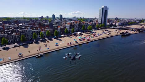 drone capturing vibrant amsterdam beach and amstel river on a sunny summer day, featuring colorful beach chairs, umbrellas, and a backdrop of traditional and modern architecture