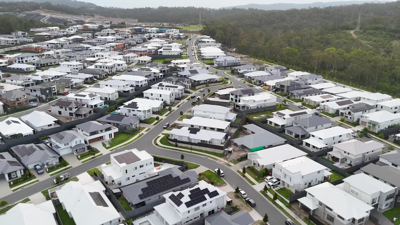 Drone glides above contemporary residential neighborhood, revealing curving streets, modern homes, and forest backdrop