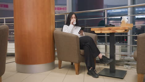 young girl sips coffee while chatting on phone in mall lounge with modern chair wooden table holding headphones tablet napkin holder city lights glow through window with soft bokeh lights