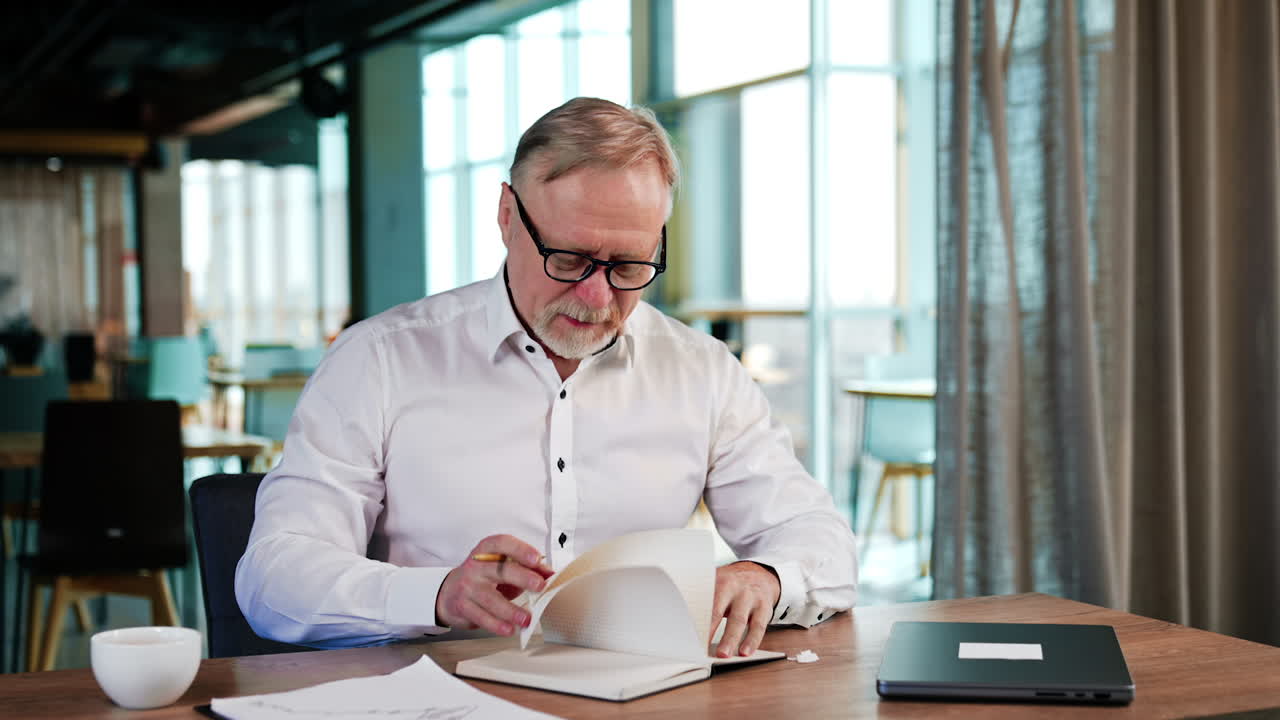 Positive Caucasian businessman sits at desk in the office. Man turns the pages of his notebook and then closes it.
