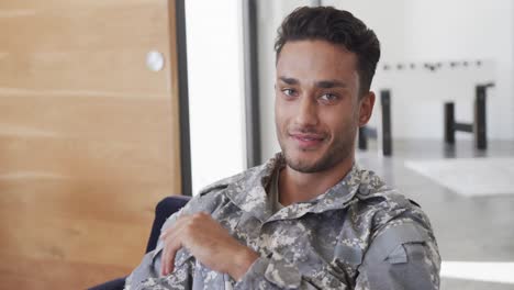 portrait of happy biracial male soldier in uniform sitting in armchair at home smiling, slow motion