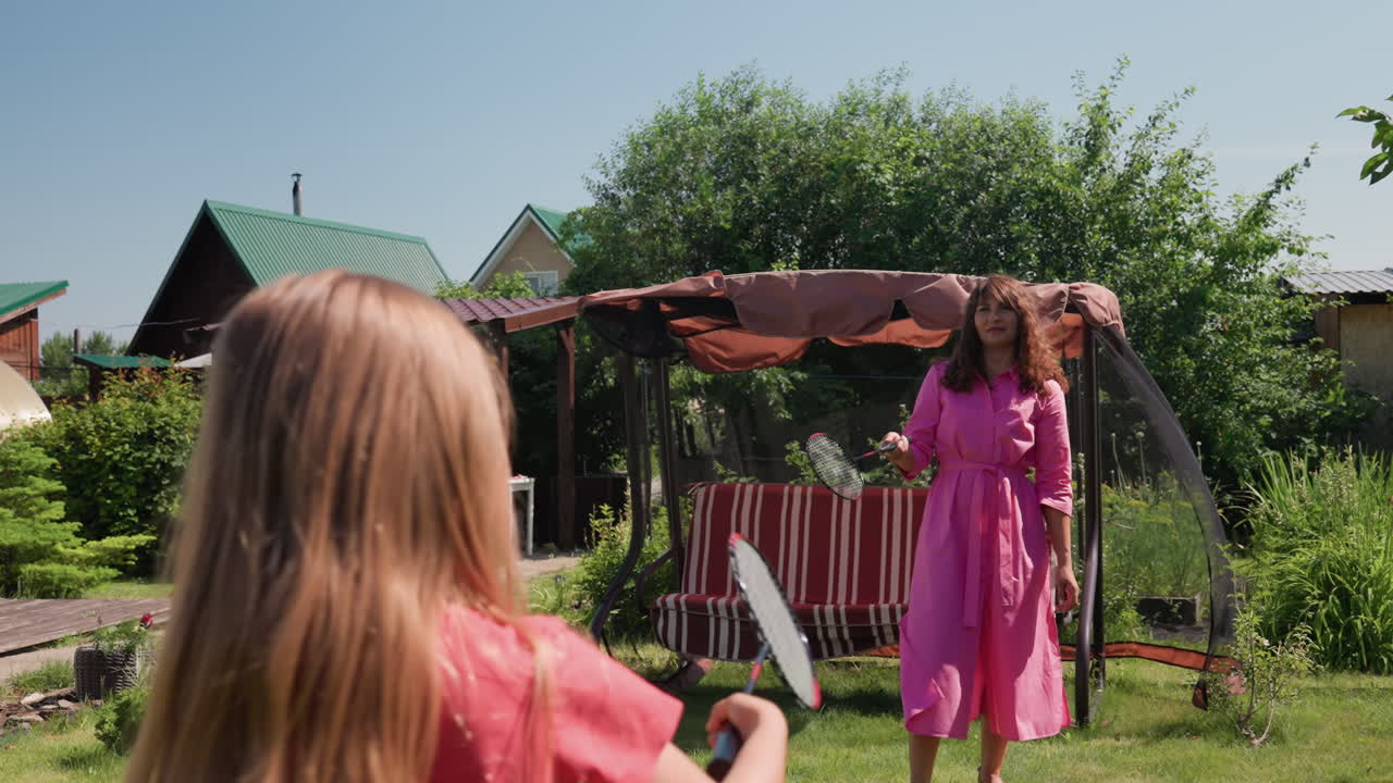 White Daughter Practices Badminton With Mother Coach In Sunny Garden, Racket And Shuttlecock Action Near Swing Canopy, Playful Instruction Tone, Energetic Backyard Sport Scene With Cottage And Green