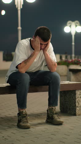 hombre cansado sentado en un banco al anochecer, profundamente en pensamiento, con luces de la calle y estructuras urbanas en el fondo, parece estresado o reflexivo en el entorno de la ciudad por la noche