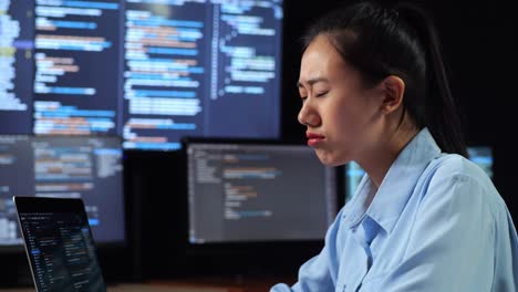 close up side view of asian female programmer yawning while writing code by a laptop using multiple monitors showing database on terminal window desktops in the office