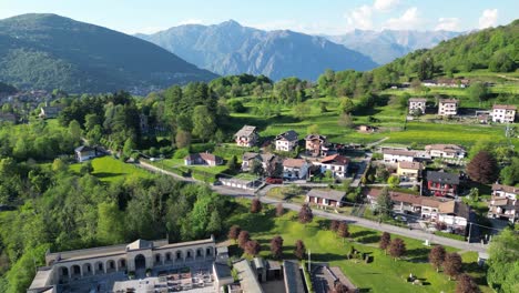 a drone slowly flies backward over san fedele near the city of como, passing above a peaceful cemetery, with rooftops, hills, and mountains glowing in the summer sun