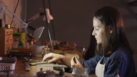 Profile-of-focused-caucasian-female-jeweller-sitting-at-desk,-making-jewelry-in-workshop