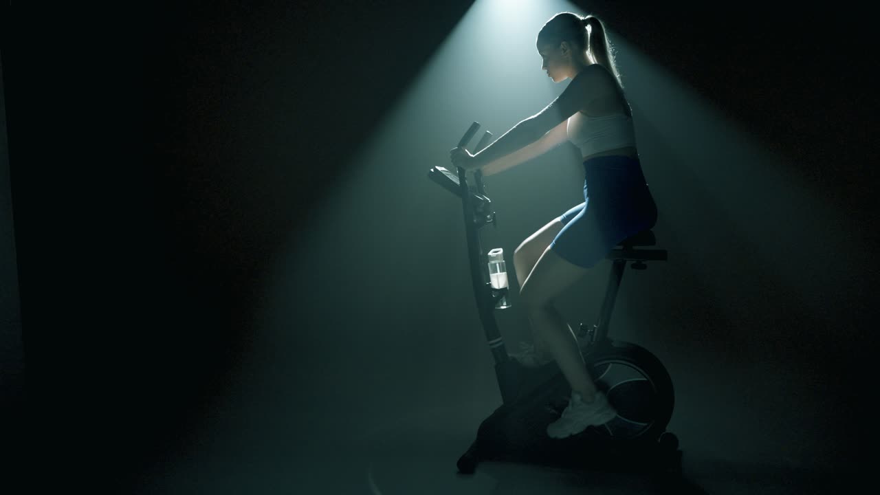 Moody cinematic shot of an empty stationary bike glowing under a spotlight in a dark studio. A woman steps from the shadows, mounts the bike, and starts pedaling with purpose and smooth motion