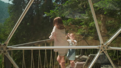 mother poses for photograph taken by daughter with camera standing on balcony overlooking dense forest in sunny summer weather. enjoying nature and modern apartment