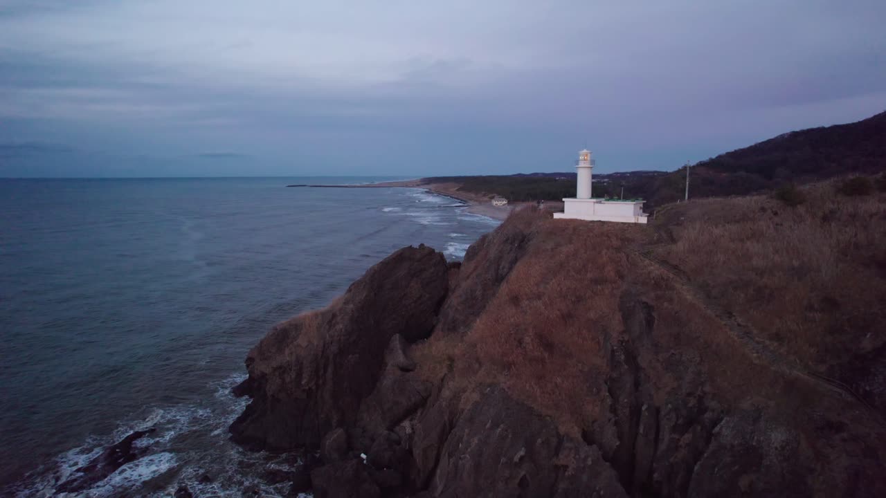 A white lighthouse overlooks the coastline of Japan, offering a serene coastal view as daylight fades