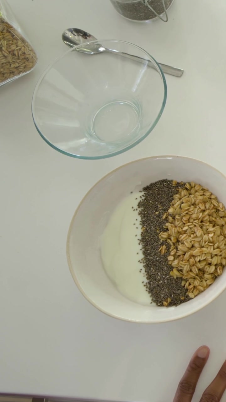Video verticale: Donna in viola che posiziona una ciotola, mescola semi di chia e avena nello yogurt sul bancone per preparare un pasto